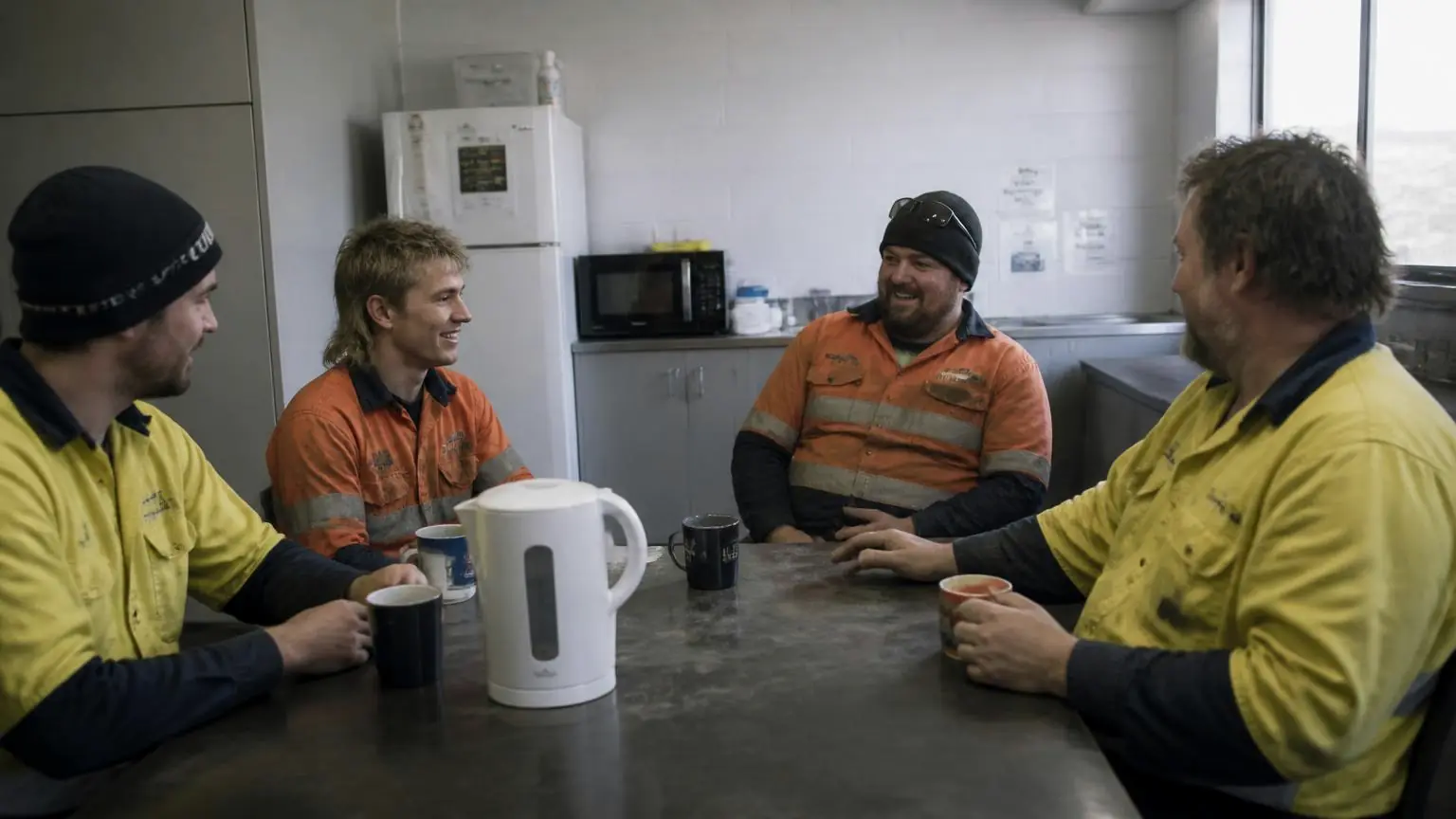 Four workshop staff in hi-vis shirts talking and laughing over mugs of tea at a staff kitchen table